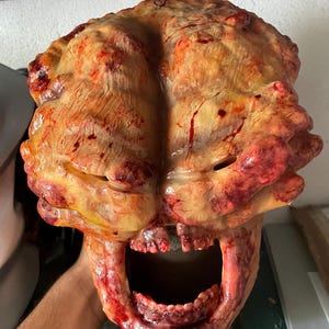 Op de afbeelding: Een masker voor het hele hoofd met een textuur die lijkt op hersenen in tinten geel, oranje en rood met dieprode bloedspatten. Het onderste gedeelte toont een open, schreeuwende mond met zichtbare tanden en rauwe, vlezige texturen.