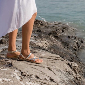 May include: A woman wearing a white skirt and silver sandals stands on a rocky shore. The water is in the background.