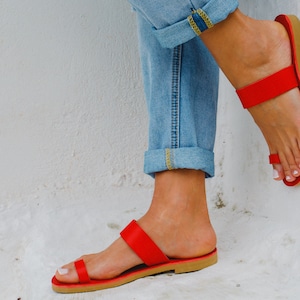 Puede incluir: Un par de sandalias de cuero rojo con tiras y un dise&ntilde;o sencillo. Las sandalias tienen dos tiras en la parte superior del pie y un bucle para los dedos. Las sandalias las lleva una persona que lleva pantalones vaqueros azules.