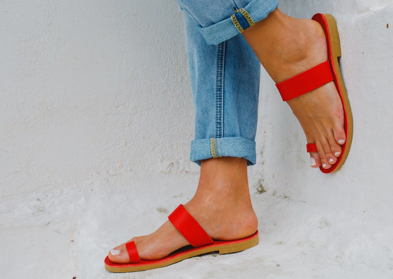 Puede incluir: Un par de sandalias de cuero rojo con tiras y un dise&ntilde;o sencillo. Las sandalias tienen dos tiras en la parte superior del pie y un bucle para los dedos. Las sandalias las lleva una persona que lleva pantalones vaqueros azules.