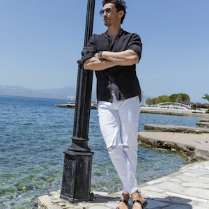 May include: A man wearing a black button-down shirt, white jeans, and black sandals stands by a lamppost near a body of water.