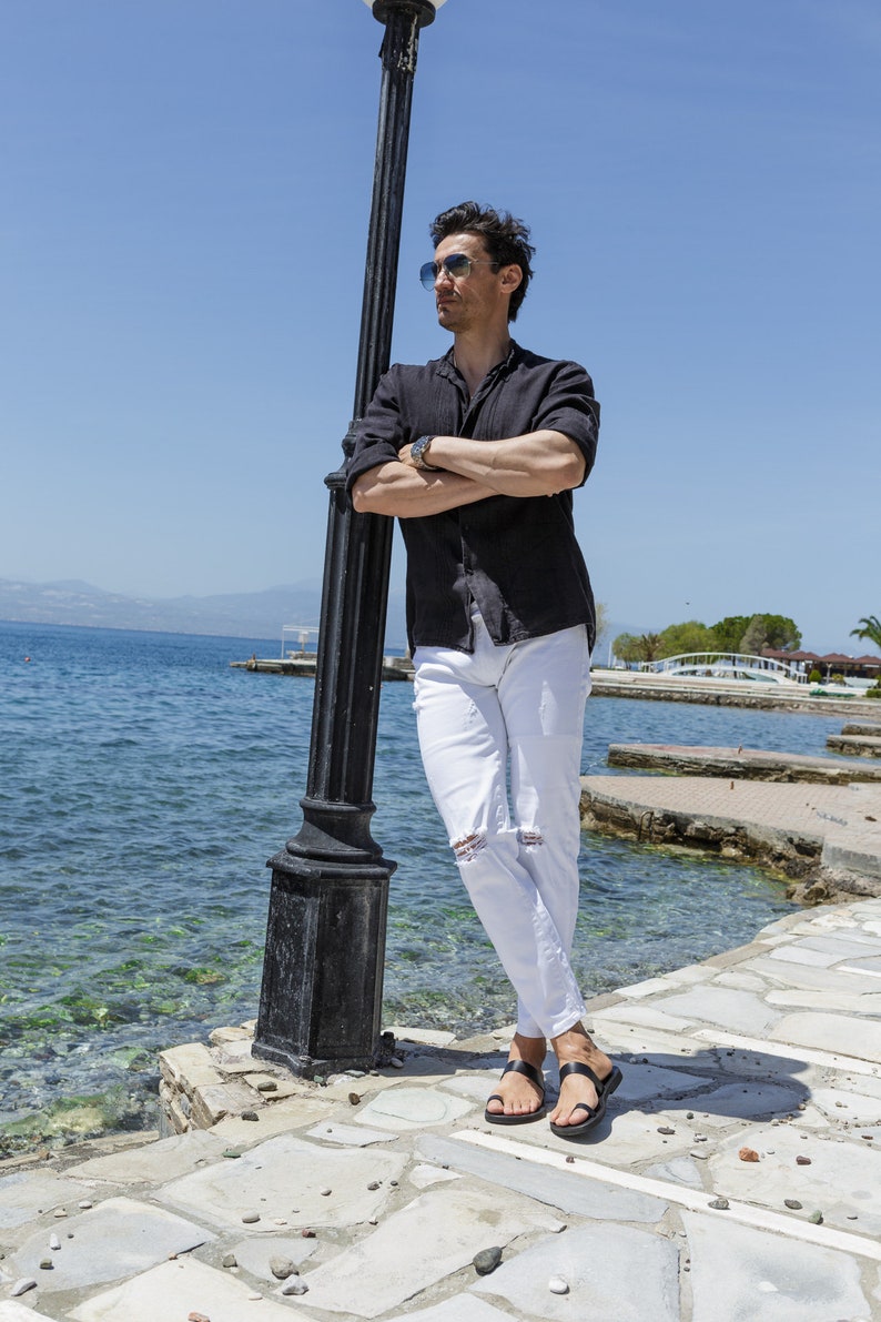May include: A man wearing a black button-down shirt, white jeans, and black sandals stands by a lamppost near a body of water.