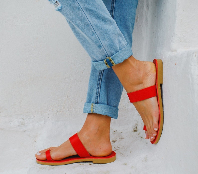 Puede incluir: Un par de sandalias de cuero rojo con una correa de dedo gordo y una correa fina de tobillo. Las sandalias las lleva una persona que lleva pantalones vaqueros azules.