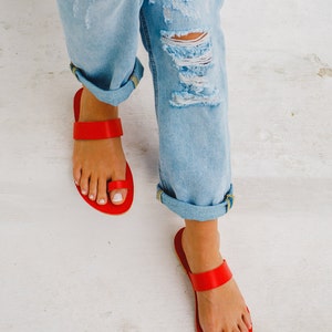 Puede incluir: Un par de sandalias de cuero rojo con una correa de aro para el dedo del pie. Las sandalias las lleva una persona que lleva pantalones vaqueros desgarrados.