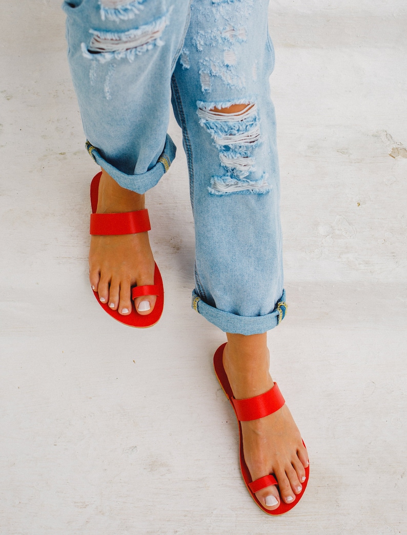 Puede incluir: Un par de sandalias de cuero rojo con una correa de aro para el dedo del pie. Las sandalias las lleva una persona que lleva pantalones vaqueros desgarrados.
