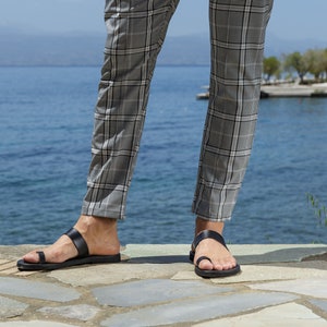 May include: A pair of black leather sandals with a toe thong and a single strap across the foot. The sandals are worn by a person standing on a stone patio.
