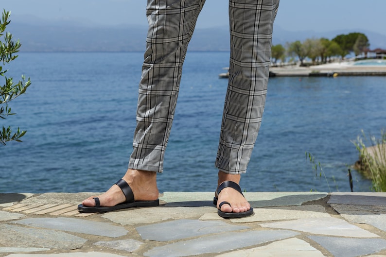 May include: A pair of black leather sandals with a toe thong and a single strap across the foot. The sandals are worn by a person standing on a stone patio.