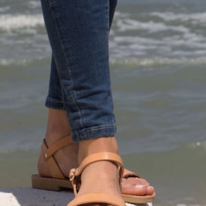 Puede incluir: Un par de sandalias de cuero marr&oacute;n con correas de tobillo. Las sandalias son usadas por una persona de pie sobre una superficie blanca con un cuerpo de agua en el fondo.