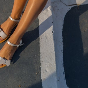 May include: A pair of white leather sandals with a bow detail on the front strap. The sandals are worn on a person's feet.