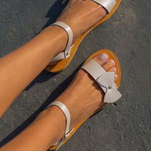 May include: White leather sandals with a bow detail on the straps. The sandals have a brown leather sole and are worn on a person's feet.