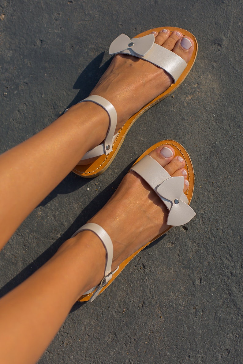 May include: White leather sandals with a bow detail on the straps. The sandals have a brown leather sole and are worn on a person's feet.