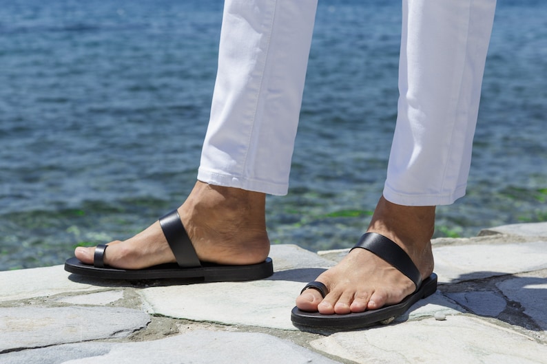 May include: A pair of black leather sandals with a toe loop and a single strap across the foot. The sandals are worn by a person standing on a stone surface.