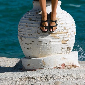 May include: A white ceramic pot with a textured surface sits on a sandy beach. The pot has a crack running down the side. The water is blue and there are whitecaps in the distance.
