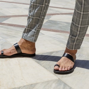 May include: A pair of black leather sandals with a toe loop and two straps. The sandals are worn on a person's feet, and the person is wearing plaid pants.