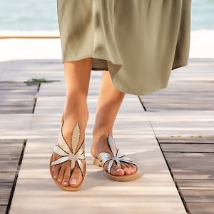 Op de afbeelding: Een paar gouden leren sandalen met een bladerenontwerp. De sandalen hebben een zwarte omtrek en worden gedragen door iemand die op een houten dek loopt.