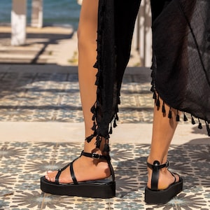 May include: Black platform sandals with multiple straps and a buckle closure. The sandals have a thick sole and are worn with a black garment with tassel trim. The sandals are on a patterned tile floor.