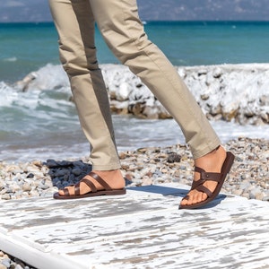 May include: A person wearing brown leather sandals with straps and buckles, walking on a white wooden surface. The sandals have a brown leather sole and are worn with beige pants.