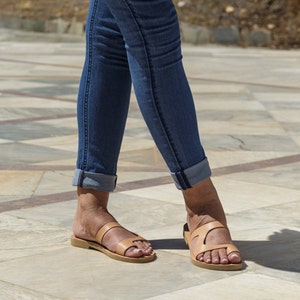 May include: A pair of tan leather sandals with two straps across the foot. The sandals are worn by a person wearing blue jeans.