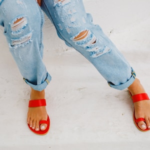 Puede incluir: Un par de sandalias de cuero rojo con una correa de anillo para el dedo del pie. Las sandalias las lleva puesta una persona que lleva pantalones vaqueros desgarrados.