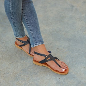 May include: A pair of dark blue leather sandals with a t-strap design and a brown sole. The sandals are worn by a person wearing blue jeans.