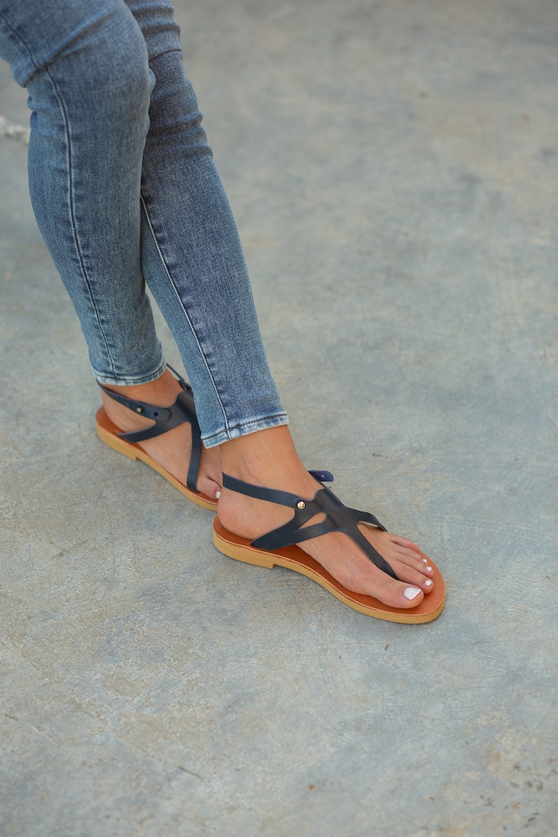 May include: A pair of dark blue leather sandals with a t-strap design and a brown sole. The sandals are worn by a person wearing blue jeans.