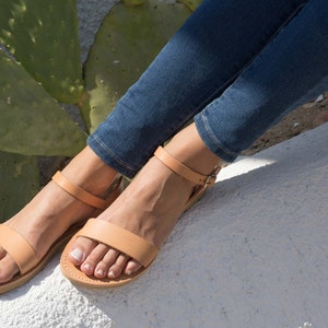 Puede incluir: Un par de sandalias de cuero marr&oacute;n con correas de tobillo. Las sandalias las lleva una persona que lleva pantalones vaqueros azules.