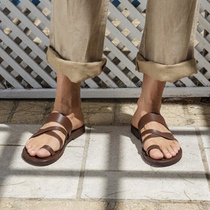 Pode incluir: Sand&aacute;lias de couro marrom com v&aacute;rias tiras e um anel para o dedo do p&eacute;. As sand&aacute;lias s&atilde;o usadas por uma pessoa em p&eacute; em um piso de ladrilhos.