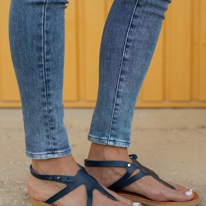 May include: A pair of dark blue leather sandals with a t-strap design and a brown sole. The sandals are worn by a person wearing blue jeans.