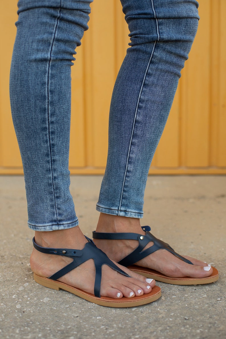 May include: A pair of dark blue leather sandals with a t-strap design and a brown sole. The sandals are worn by a person wearing blue jeans.
