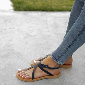 May include: A pair of black leather sandals with a strap across the foot and a buckle closure. The sandals have a brown leather sole and are worn by a person with bare feet.