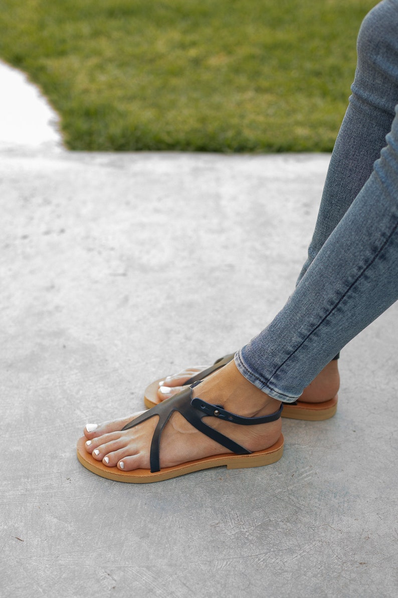 May include: A pair of black leather sandals with a strap across the foot and a buckle closure. The sandals have a brown leather sole and are worn by a person with bare feet.
