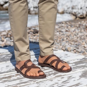 May include: A pair of brown leather sandals with multiple straps. The sandals are worn by a person standing on a white wooden surface.