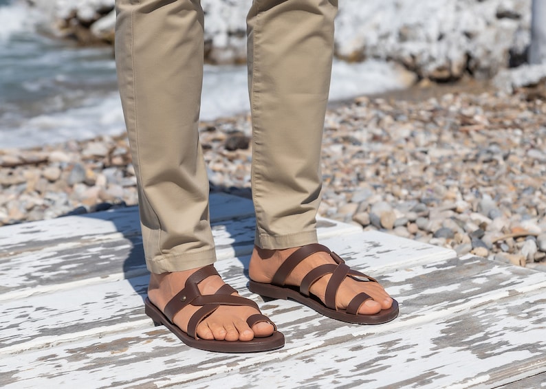 May include: A pair of brown leather sandals with multiple straps. The sandals are worn by a person standing on a white wooden surface.