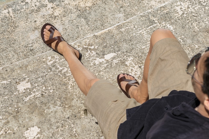 May include: A person wearing brown leather sandals with straps, khaki shorts, and a black shirt. The person is sitting on a concrete surface.