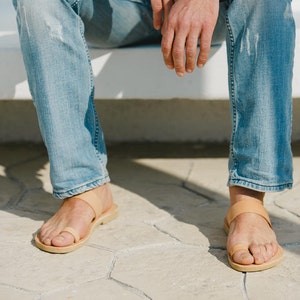 May include: A pair of tan leather sandals with a thong strap and a single strap across the foot. The sandals are worn by a person wearing blue jeans.