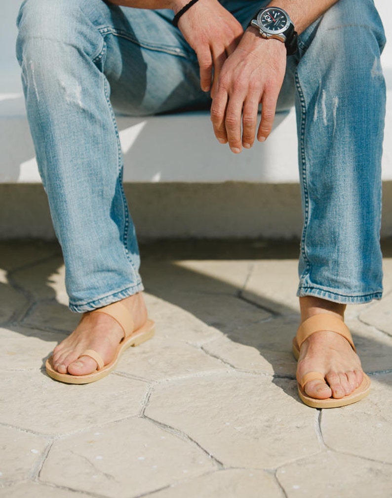 May include: A pair of tan leather sandals with a thong strap and a single strap across the foot. The sandals are worn by a person wearing blue jeans.