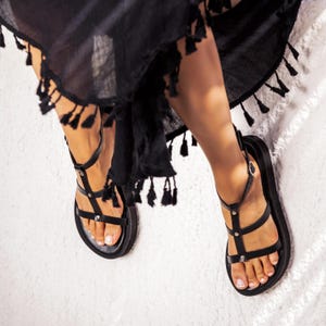 May include: Black leather sandals with multiple straps and silver-tone studs. The sandals have a flat sole and a buckle closure. The image shows the sandals on a white surface, with a black garment with tassels in the background.