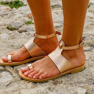 May include: A pair of gold leather sandals with a toe ring and ankle strap. The sandals are on a rocky surface.