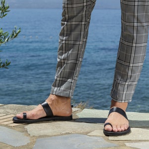May include: A pair of black leather sandals with a toe thong strap. The sandals are worn by a person standing on a stone path with a body of water in the background.