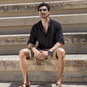 May include: A man wearing sunglasses and a black linen shirt sits on concrete steps wearing brown leather sandals.