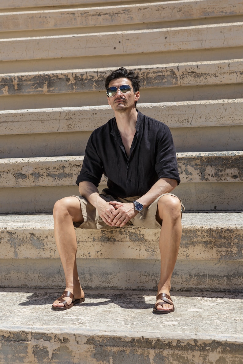 May include: A man wearing sunglasses and a black linen shirt sits on concrete steps wearing brown leather sandals.