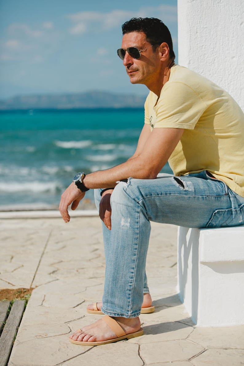 May include: A man wearing sunglasses, a yellow t-shirt, blue jeans, and brown leather sandals sits on a white ledge by the ocean.