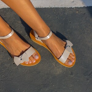 May include: A pair of white leather sandals with a bow detail on the toe strap. The sandals have a brown leather sole.