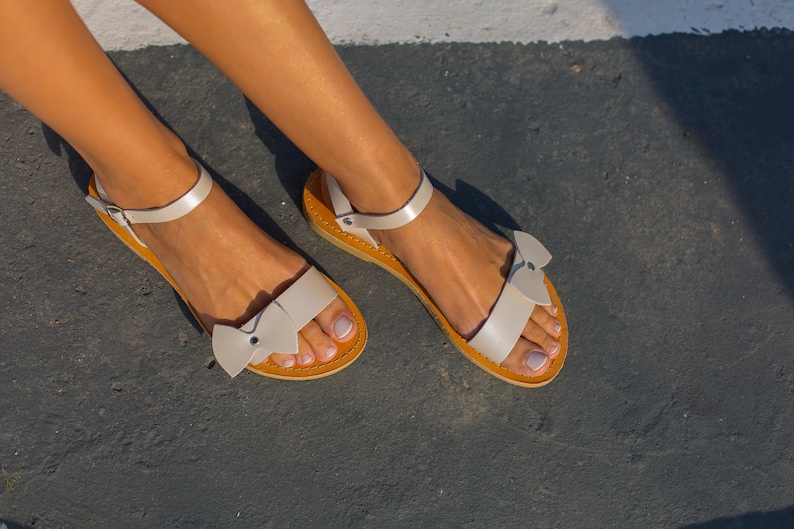 May include: A pair of white leather sandals with a bow detail on the toe strap. The sandals have a brown leather sole.