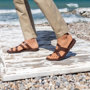 May include: Brown leather sandals with multiple straps and a buckle on the foot strap. The sandals are worn by a person standing on a weathered white wooden surface.