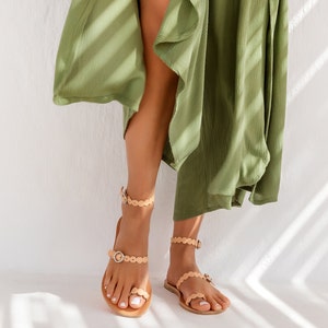 Puede incluir: Un par de sandalias de cuero color canela con un detalle de borde festoneado y un cierre de hebilla. Las sandalias las lleva una persona que lleva un vestido verde.