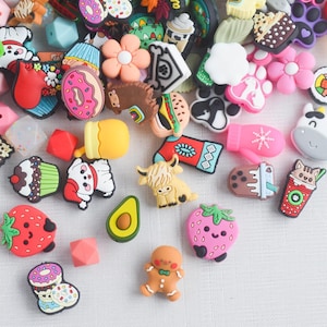 May include: A collection of colorful shoe charms in various shapes, including food items like donuts, cupcakes, and strawberries, along with flowers, animals, and other decorative designs. The charms are made of rubber and have a flat back.