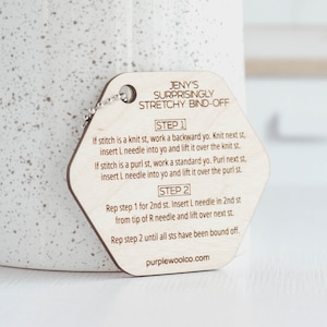 May include: A wooden hexagonal tag with instructions for a stretchy bind-off knitting technique. The tag is light brown with dark brown text. The text reads "JENY'S SURPRISINGLY STRETCHY BIND-OFF" and includes step-by-step instructions for knitting. The tag has a small metal ring at the top.