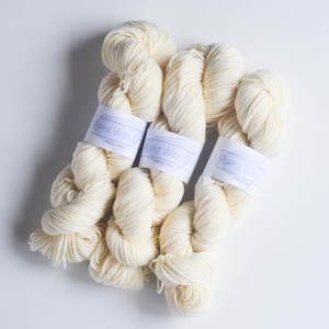 May include: Three skeins of undyed yarn are displayed against a white background. Each skein is wrapped with a white label that reads "Purple Wool Co." in a purple font. The yarn is a creamy off-white color, and the texture appears soft and fluffy.