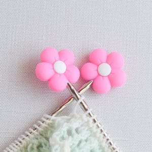 May include: Two pink flower-shaped stitch markers with white centers rest near knitting needles and a partially completed light green and white knitted project. The background is a white, textured fabric.